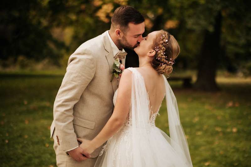 Das Brautpaar hält sich bei den Händen und küsst sich zart auf einer grünen Wiese mit erstem Herbstlaub